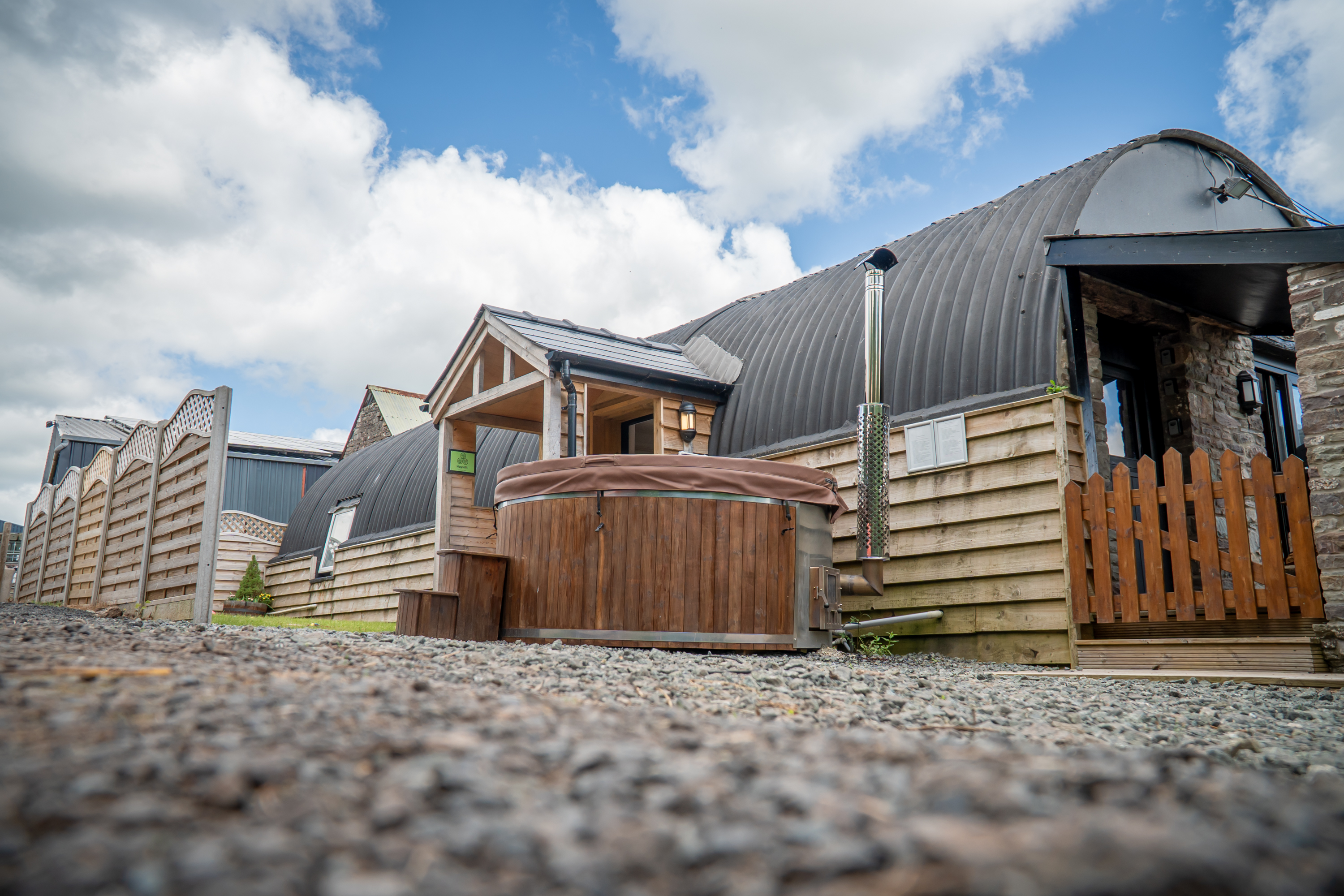 An image of a hayloft with a tin Barrell Vault roof and exterior wooden cladding. Converted into accommodation with an entrance porch, outdoor wooden fired hot tub and a small lawned area for outdoor games.