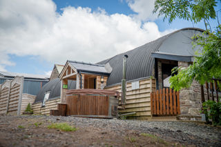 An image of a hayloft with a tin Barrell Vault roof and exterior wooden cladding. Converted into accommodation with an entrance porch, outdoor wooden fired hot tub and a small lawned area for outdoor games.